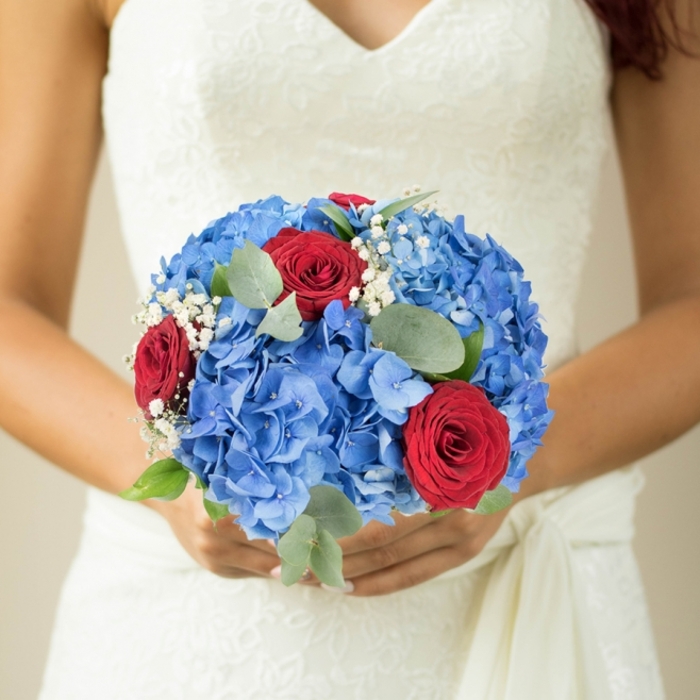 Every element of this hand-tied bouquet is chosen and placed for harmony and meaning, with a confident eye for texture and contrast. The blue hydrangeas are full and generous-tones of cerulean and pale sky that suggest calm seas and a promise of gentle days ahead-forming a voluminous base that cushions the composition. Scarlet red roses are nestled deliberately among the blues, their layered petals curling softly as if holding private stories of love; their rich colour provides a thrilling counterpoint that animates the whole arrangement. Tiny clusters of gypsophila float between the larger blooms, introducing a sense of weightlessness and innocent sparkle akin to first snow or confetti. Eucalyptus leaves, with a quiet matte sheen, braid through the bouquet, offering a woodland freshness and a subtle fragrance that grounds the bouquet in the senses. The arrangement reads beautifully in natural daylight-whether captured near the arches of St John's Gate or beside a doorway on Farringdon Road-its colours appearing saturated yet comforting. Designed to be carried down the aisle or given as a deeply personal gift, the bouquet is at once a promise and a celebration: fragrant, tactile, and composed with the considered touch of a florist who understands ceremony, place, and feeling. It is a compact, evocative composition meant to be admired closely and remembered for its colour, scent, and emotional clarity.